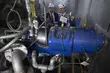 Two engineers wearing protective helmets inspecting a large blue industrial machine surrounded by pipes and gauges.